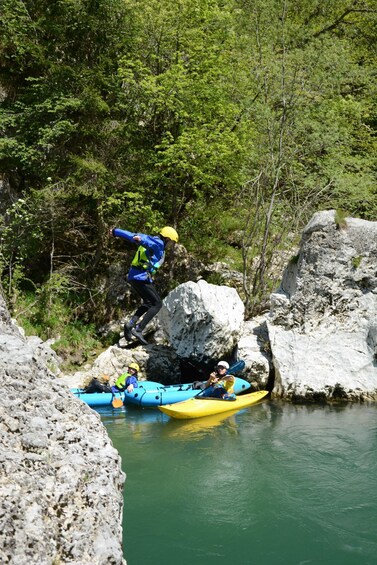 A person in a blue jacket and yellow helmet jumping from a rock into a river, with two kayaks and another person in the water. 2