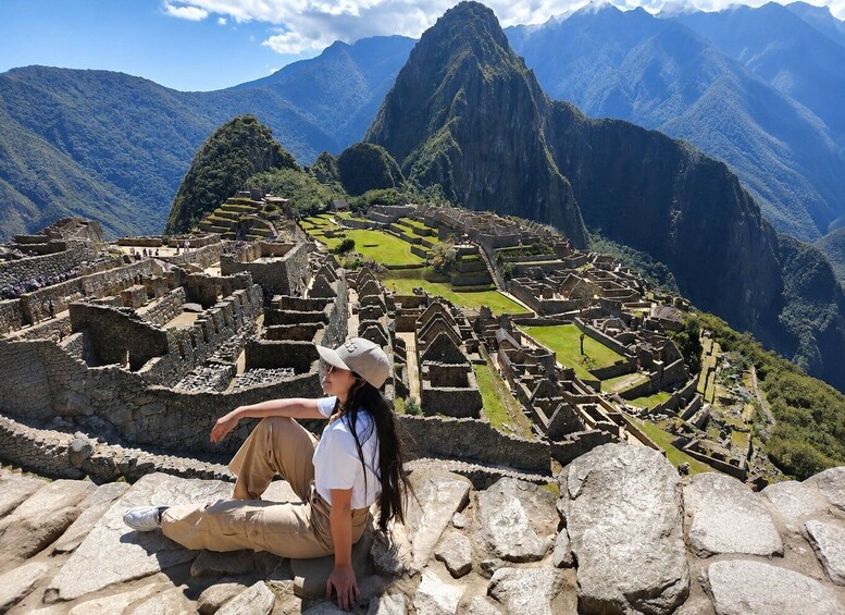 A person sitting on a stone ledge overlooking Machu Picchu, with the iconic Incan ruins and surrounding mountains in the background. 3
