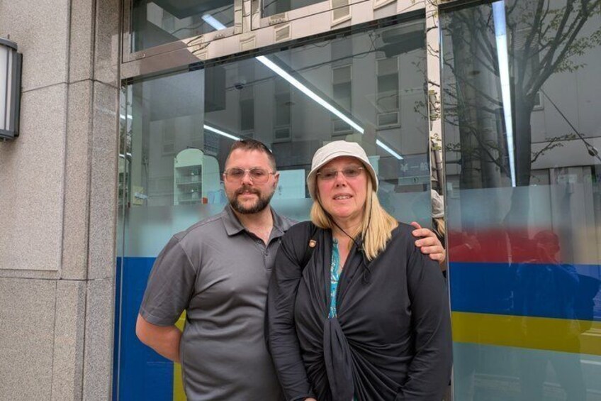 Two people standing in front of a building with a glass facade and a blue and yellow stripe. 2