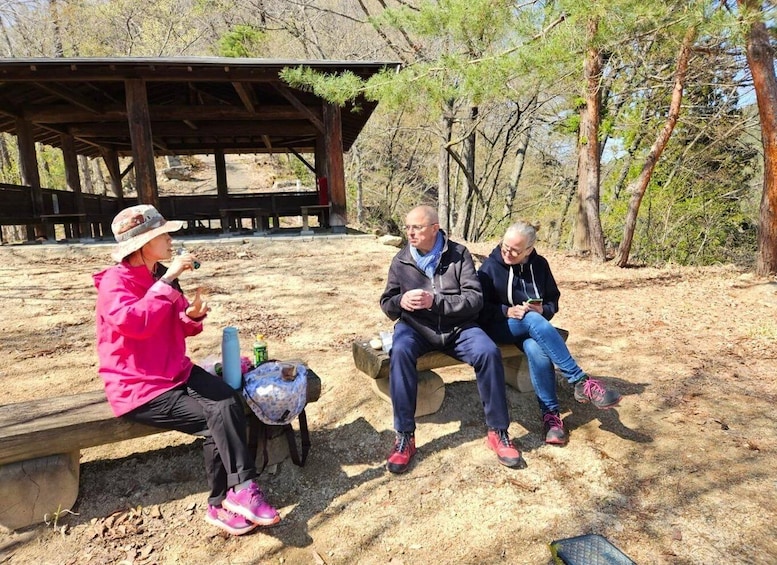 Three people sitting on a bench outdoors with a wooden structure in the background. 3