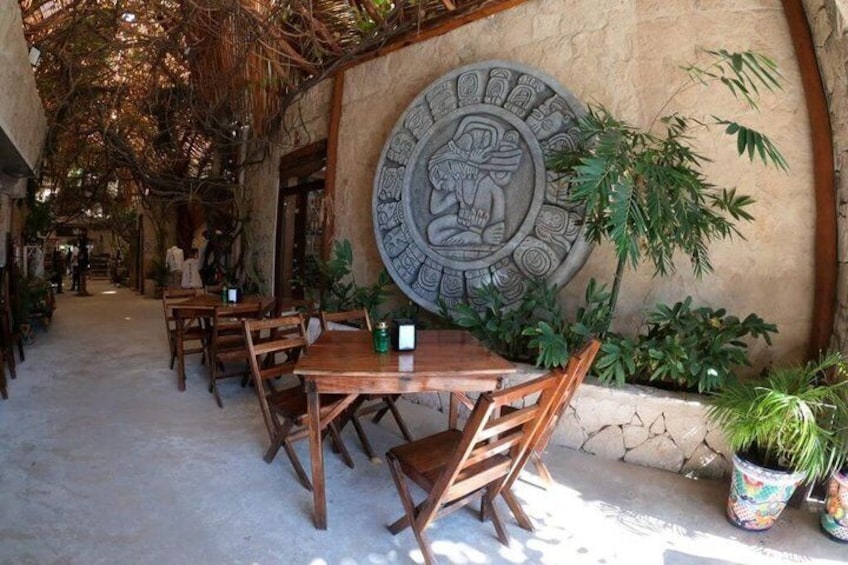 An outdoor dining area with wooden tables and chairs, surrounded by plants and a large stone wall with an ancient carving. 4