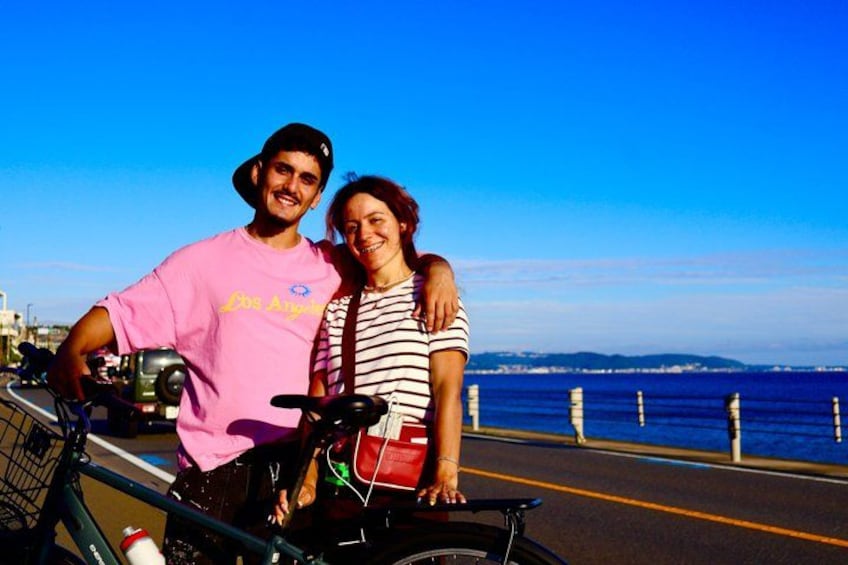 Two people standing next to a bicycle on a coastal road with a clear blue sky and a view of the ocean. 1