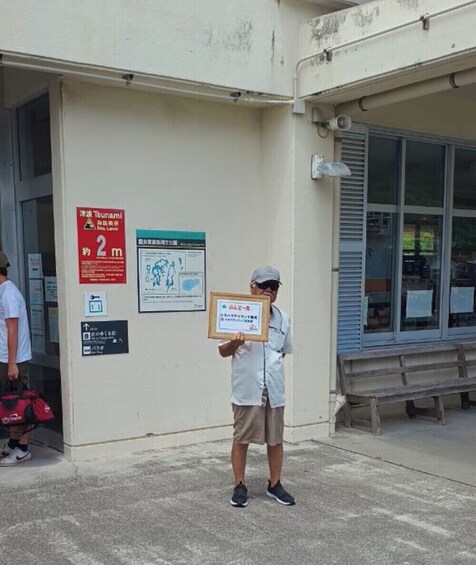 A person holding a framed certificate in front of a building with a tsunami warning sign. 2