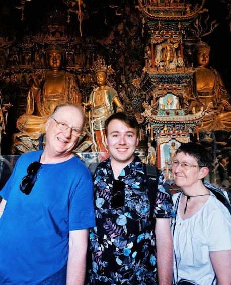 Three people standing in front of a large golden statue with intricate designs and smaller statues in the background. 2