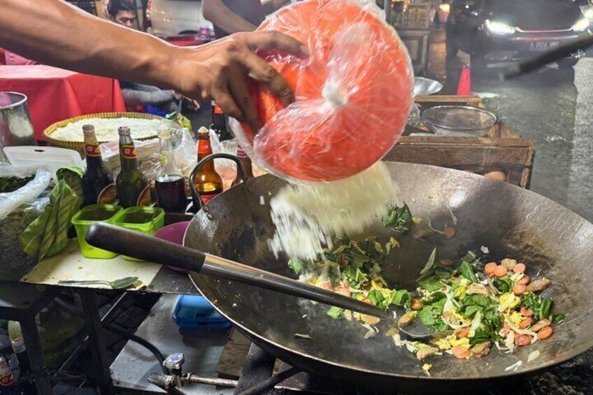 A wok with vegetables and a person pouring a liquid from a plastic bag. 5