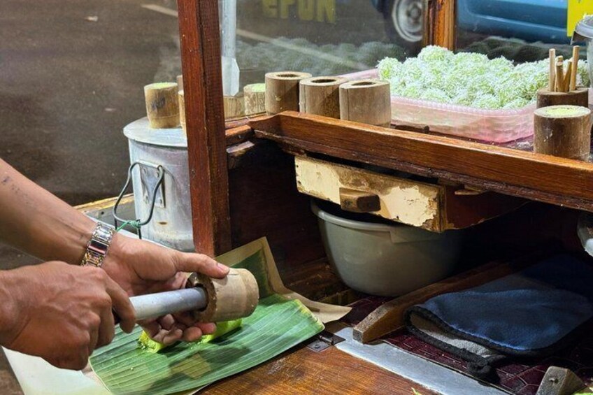A person using a manual tool on a green leaf, possibly for food preparation, with a wooden table and various containers in the background. 3