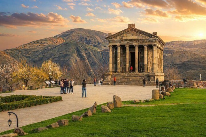 A classical building with columns, surrounded by a grassy area and a paved walkway, with mountains in the background. 4