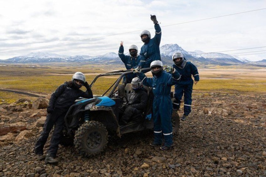 A group of people in blue overalls and helmets around a blue off-road vehicle on a rocky terrain with mountains in the background. 2