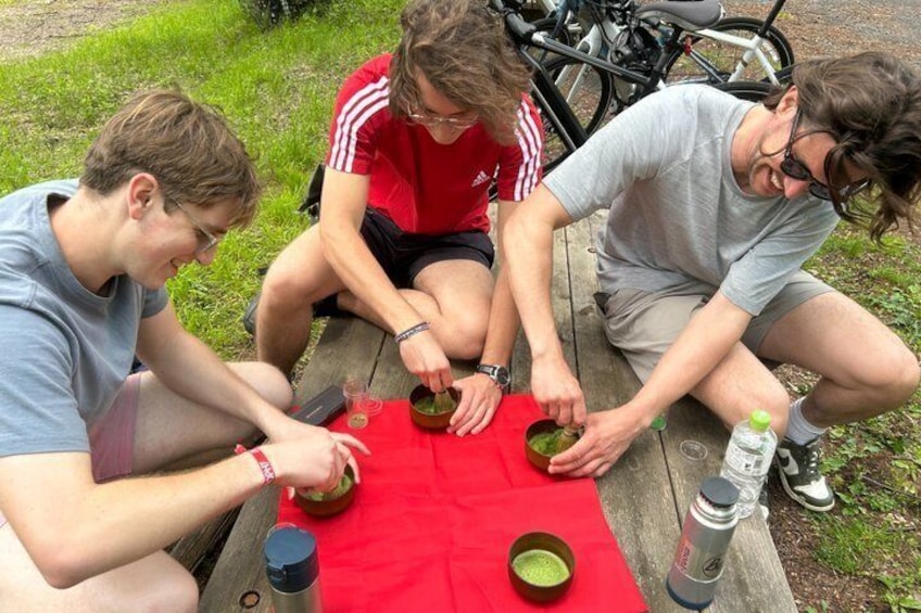 Three people sitting at a picnic table outdoors with bicycles in the background. 1