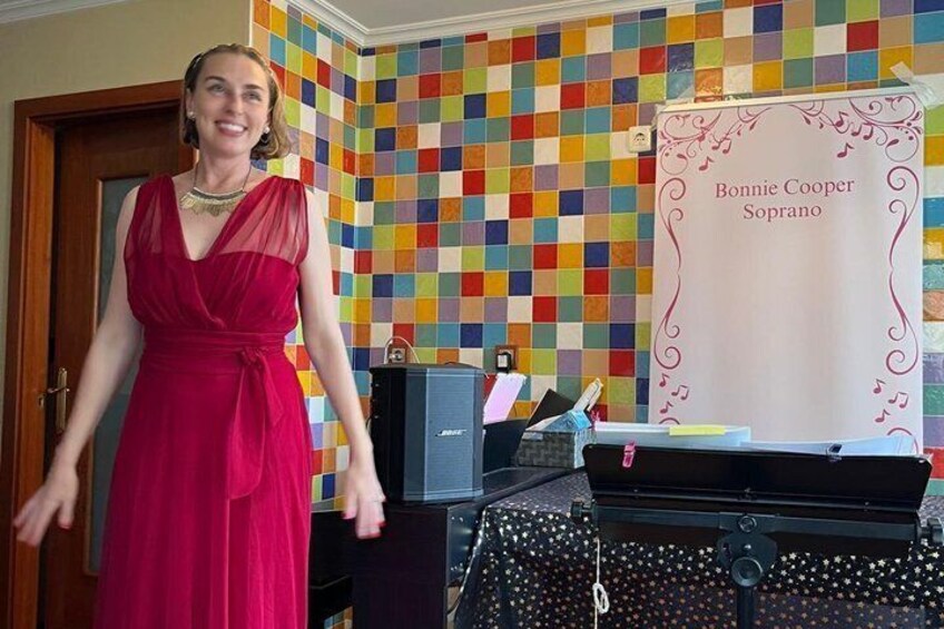 A woman in a red dress standing in front of a colorful checkered wall, with a sign reading 'Bonnie Cooper Soprano' and a piano in the background. 5