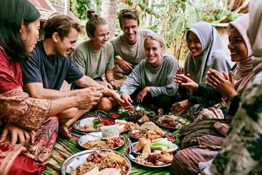 A group of people sitting around a table with various dishes, in a lush, green outdoor setting. 1