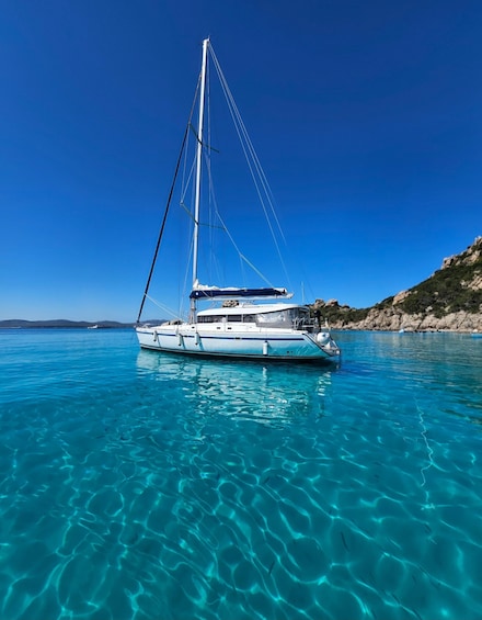 A sailboat on clear turquoise water with a rocky coastline in the background. 1