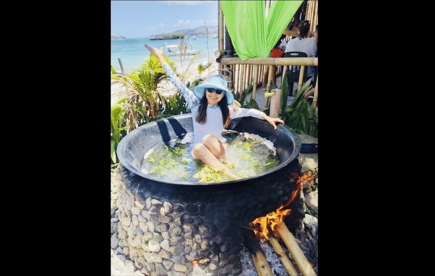 A person sitting in a large black bowl filled with water, surrounded by rocks and a fire. 3