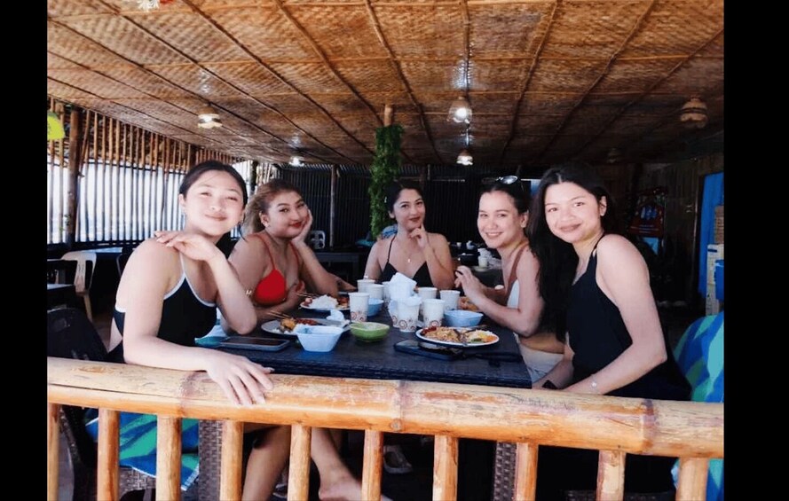 A group of people sitting at a table with food and drinks, in a rustic setting with a bamboo ceiling. 2
