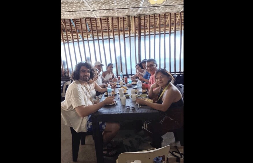 A group of people sitting at a table with food and drinks, under a thatched roof structure. 4