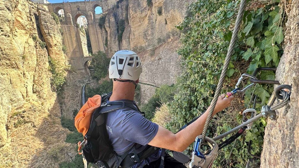 A person rappelling down a rocky cliff with a helmet and harness, overlooking a canyon with ancient bridges. 1