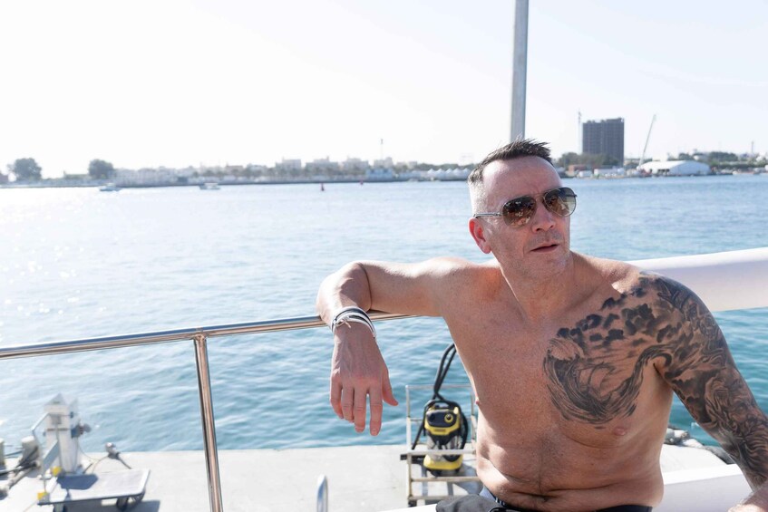 A man with tattoos on his arm leaning on a boat railing, with a water body and distant buildings in the background. 3