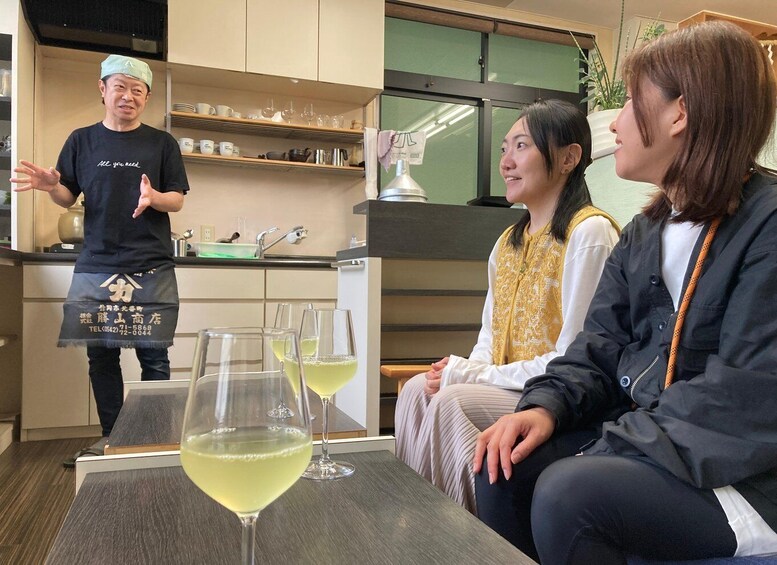 A person in a black apron with text, standing in a kitchen with shelves and a sink. There are three glasses of a yellowish drink on a table in the foreground. 1