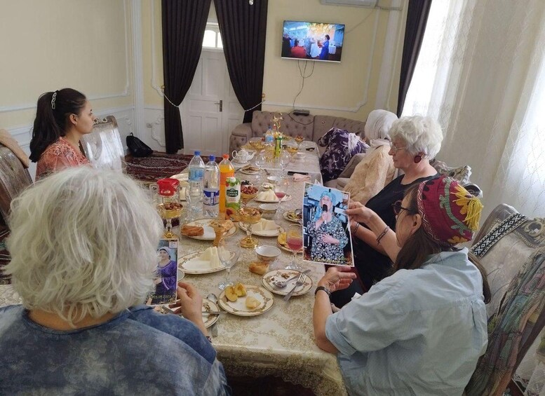 A group of people sitting around a table with food and drinks, a television mounted on the wall, and a window with curtains. 4