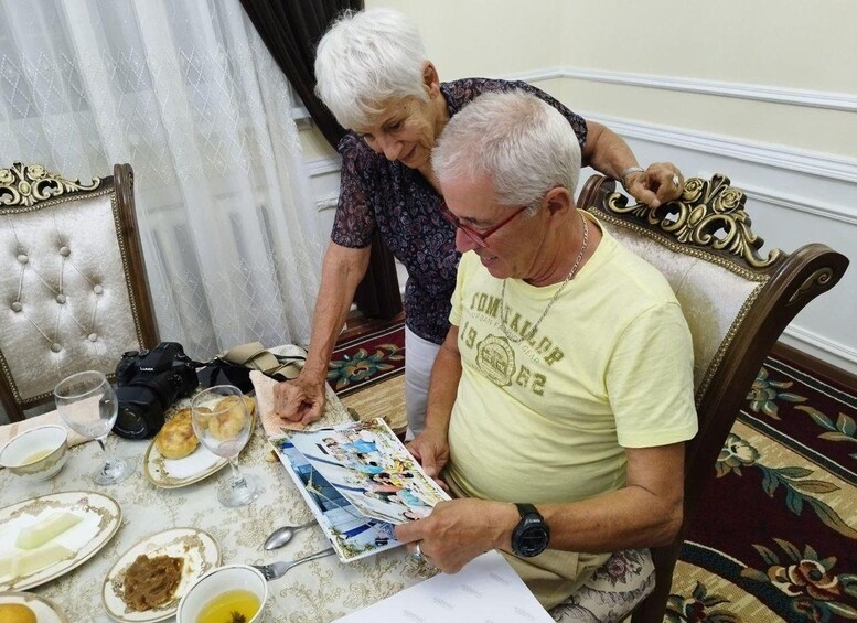 An elderly man and woman at a dining table with food remnants, a camera, and a photo album. 2