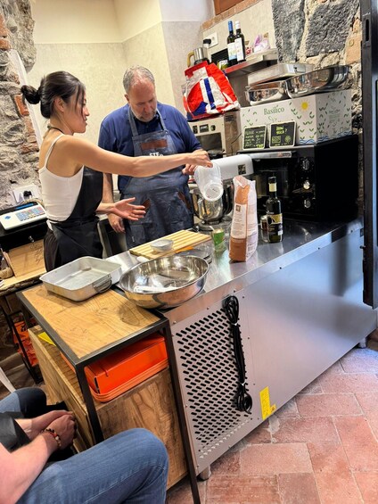 A kitchen with a person pouring liquid into a bowl, another person standing nearby, and a counter with various kitchen items. 4