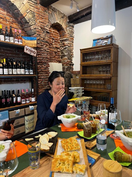 A person sitting at a table with a variety of food and drinks, including wine bottles and glasses, in a rustic setting with brick walls and wooden shelves. 2