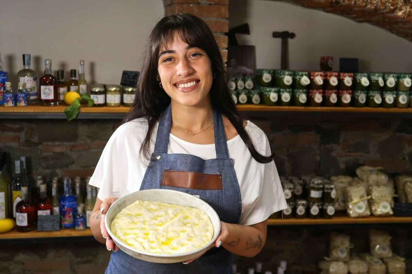 A person holding a dish with a creamy, yellowish mixture, standing in a kitchen with shelves stocked with various bottles and jars. 1