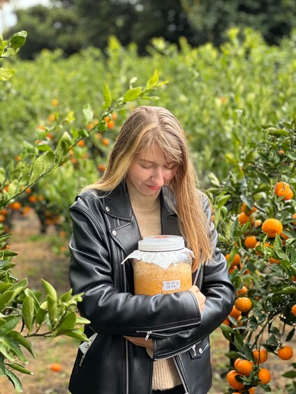 A person holding a jar of preserved food in an orchard with orange trees. 2