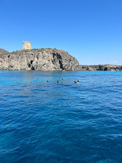 A coastal scene with a historic tower on a rocky cliff, clear blue water, and a few divers swimming. 2