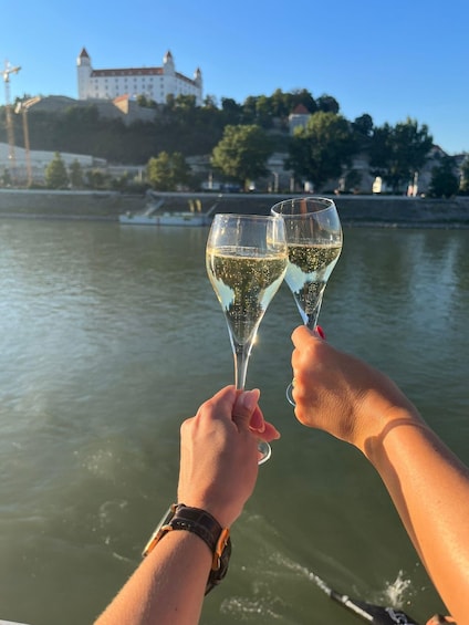 Two hands holding champagne glasses toasting, with a castle and river in the background. 1