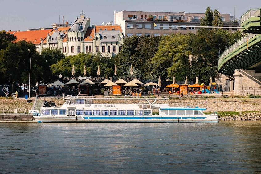 A river with a blue and white boat named LOD MARTIN. There are outdoor cafes with umbrellas along the riverbank. 2
