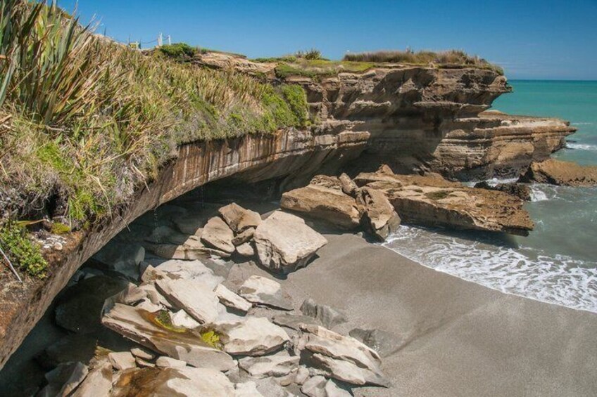 A coastal landscape with a rocky cliff, a sandy beach, and a clear blue sky. 4