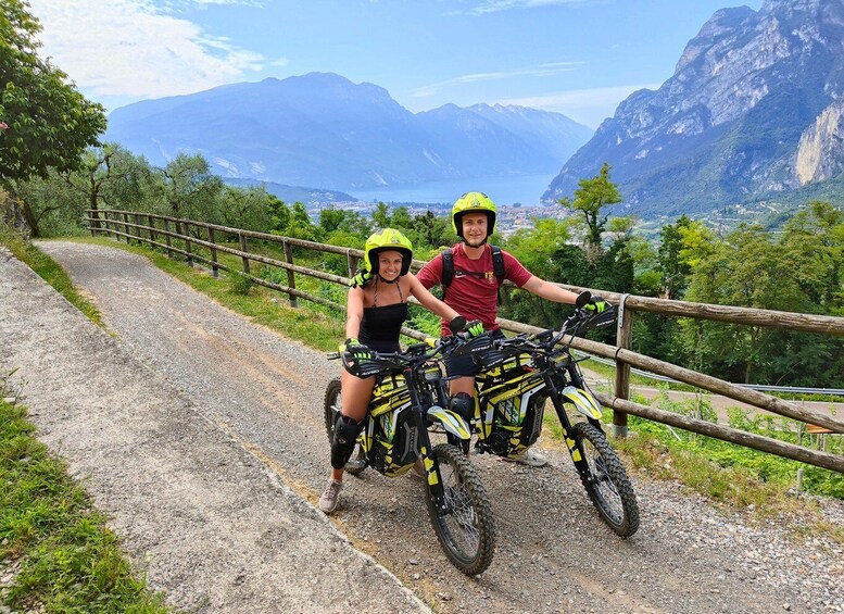 Two people on mountain bikes on a gravel path with a wooden fence, surrounded by greenery and mountains in the background. 5