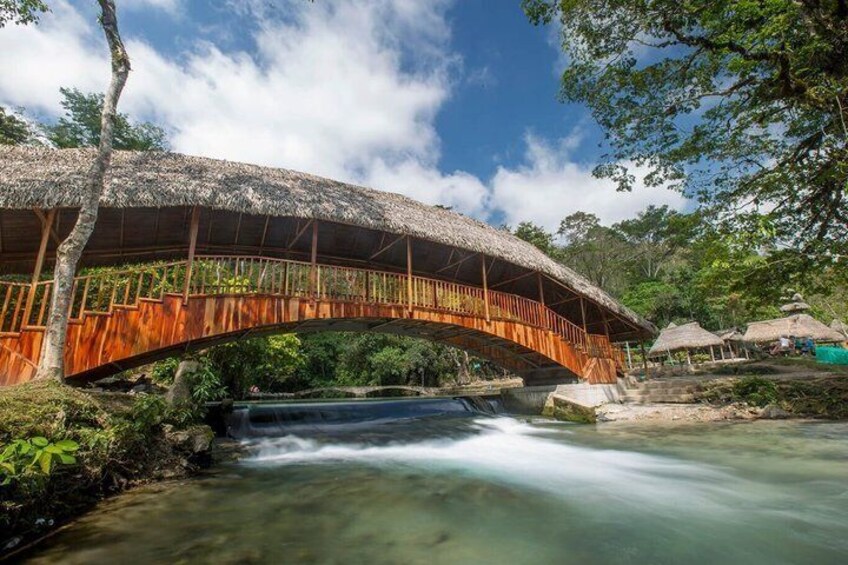 A wooden bridge over a flowing river, surrounded by lush greenery and a clear blue sky. 3