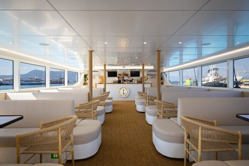 A spacious interior with white seating, a central bar area, and large windows overlooking the sea. 1