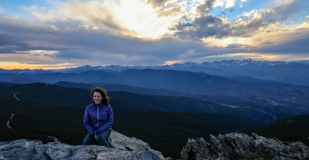 A person sitting on a rocky outcrop overlooking a mountainous landscape with a winding road and a sunset. 5