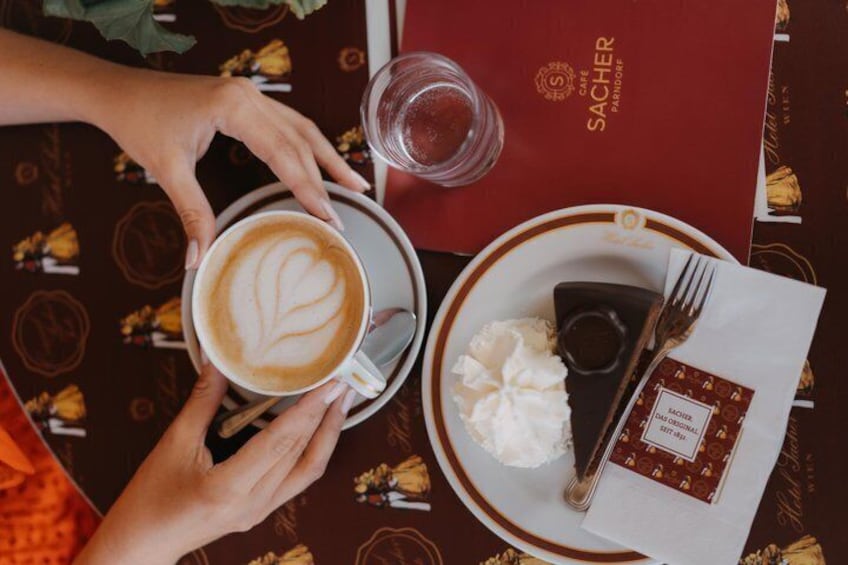 A person holding a cup of coffee with a heart-shaped latte art, a plate with whipped cream and a chocolate dessert, and a menu on a table with a patterned tablecloth. 1