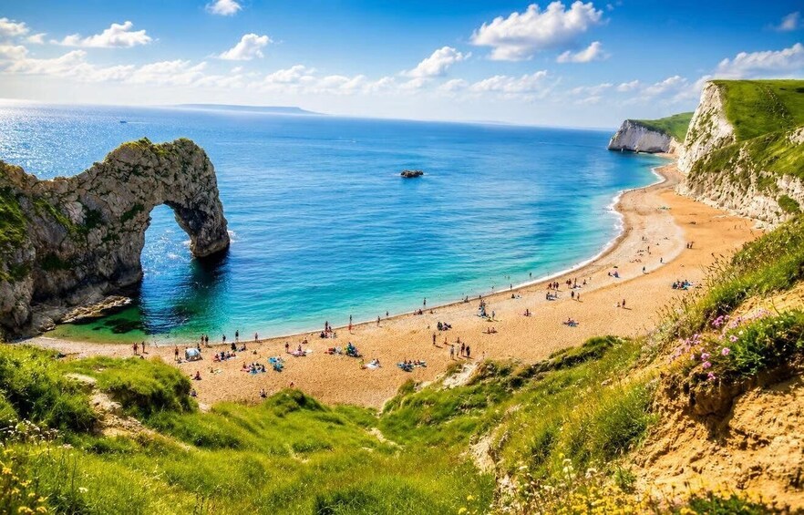 A coastal beach with a prominent rock arch formation, clear blue waters, and a sandy shore populated with people. 5