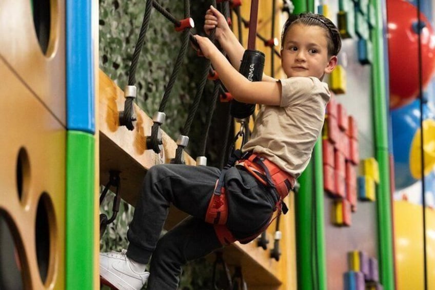 A child climbing a wooden climbing wall with colorful panels and safety harnesses. 3