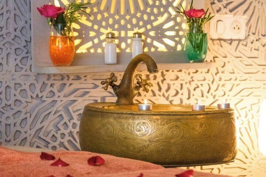A brass basin with a curved spout, a wall with intricate patterns, and two vases with flowers. 2