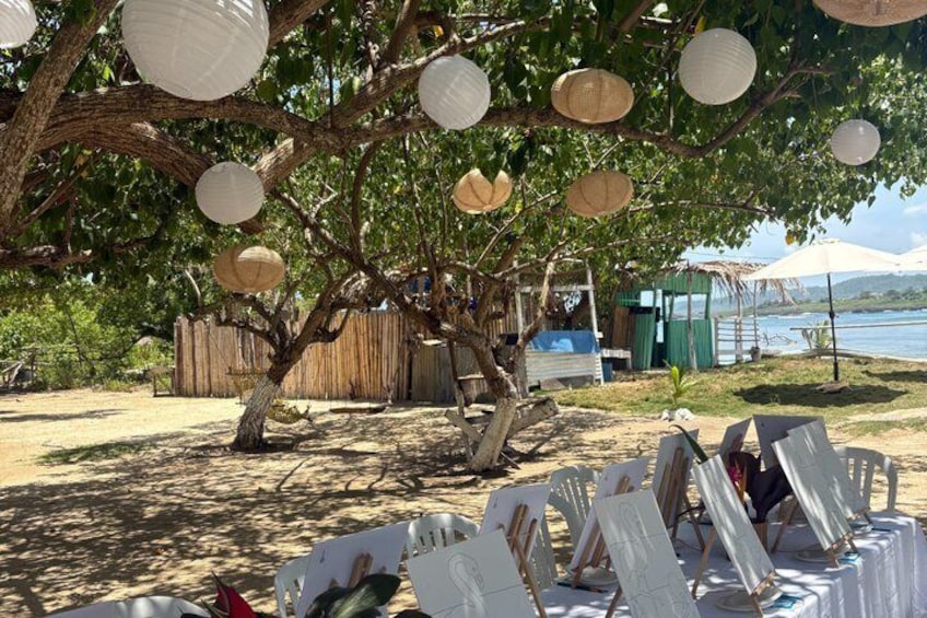 A beach setting with a tree, white paper lanterns, and easels with canvases. 4
