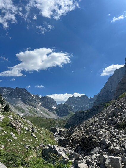 A mountainous landscape with rocky terrain, green vegetation, and a clear blue sky with clouds. 4