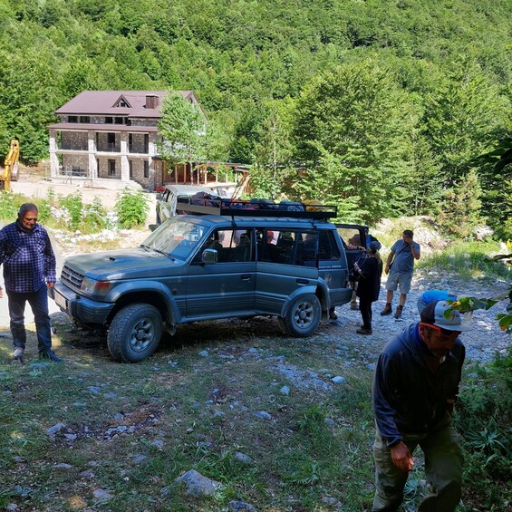 A group of people around a parked SUV in a forested area with a building in the background. 3