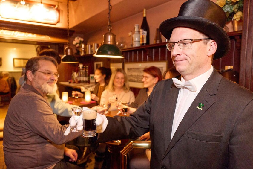 A man in a suit and hat holding a glass of beer, surrounded by people in a bar setting. 1