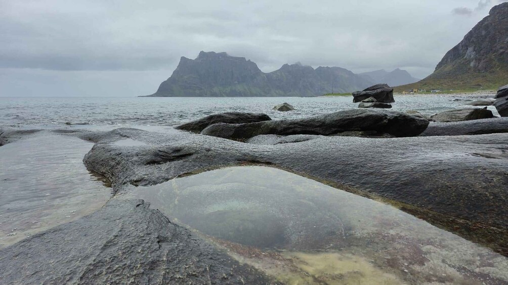 A rocky shoreline with clear water, large boulders, and distant mountains under a cloudy sky. 5