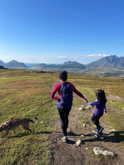 A person hiking with a child and a dog on a grassy trail with mountains in the background. 4