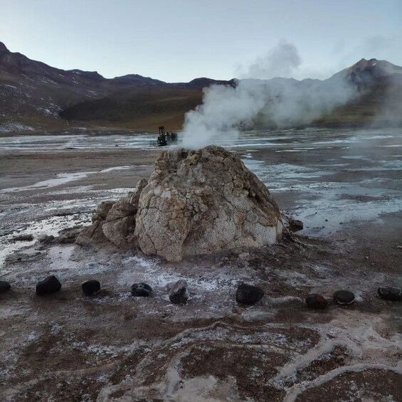A geothermal area with steam rising from the ground, surrounded by a rocky landscape and distant mountains. 1