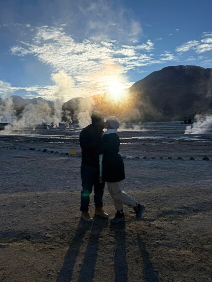 A couple embracing on a rocky shore with steam rising from the ground and a mountain in the background. 3