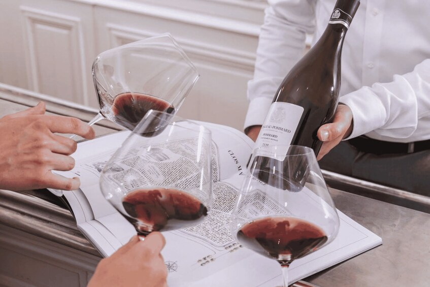 A person pouring wine into a glass, with a bottle and a newspaper on the table. 1