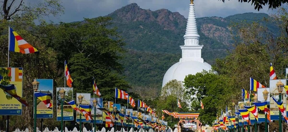 Eine weiße Stupa mit einer Spitze, umgeben von Gebetsfahnen und Wimpeln, vor einer Bergkulisse. 4